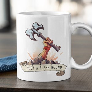 May include: White ceramic mug with a cartoon illustration of a hand holding an axe emerging from a pile of rocks. The text "Just a flesh wound" is on a banner below the image.