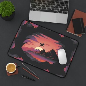 May include: A black mousepad with a dark purple, pink, and blue sunset scene with a silhouette of a dragon flying in the sky. The mousepad is on a gray surface with a laptop, a cup of coffee, and pencils.