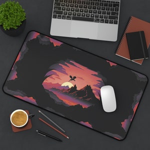 May include: A black mousepad with a colorful illustration of a dragon flying over a mountain range at sunset. The mousepad is on a black surface with a laptop, a notebook, and a cup of coffee.