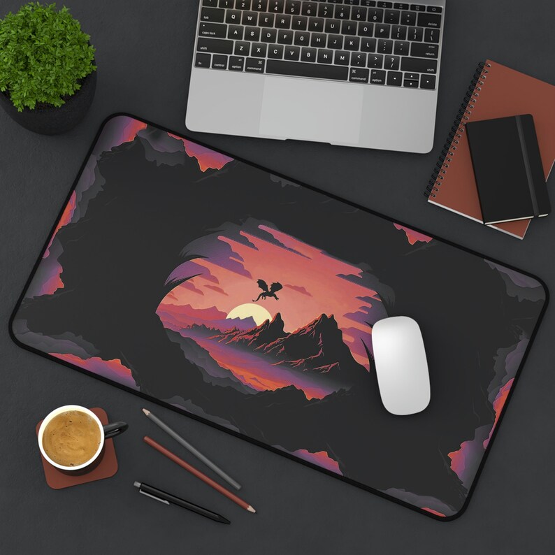 May include: A black mousepad with a colorful illustration of a dragon flying over a mountain range at sunset. The mousepad is on a black surface with a laptop, a notebook, and a cup of coffee.