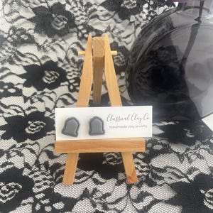 May include: A pair of gray clay earrings shaped like tombstones. The earrings are displayed on a small wooden easel with a white card that reads "Classical Clay Co. handmade clay jewelry".