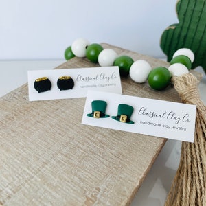 May include: Two pairs of handmade clay earrings. One pair features a black pot of gold with gold glitter. The other pair features green leprechaun hats with gold buckles. The earrings are displayed on a wooden surface with a sign that reads "Classical Clay Co. handmade clay jewelry".