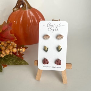 Puede incluir: Un conjunto de tres pares de pendientes de arcilla hechos a mano con un diseño de calabaza, maíz y pastel. Los pendientes se muestran en un pequeño caballete con el texto "Classical Clay Co. handmade clay jewelry".