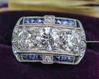 May include: A silver ring with three large diamonds and a band of blue sapphires. The ring is in a purple velvet ring box.