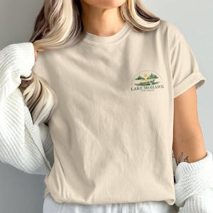 May include: Beige t-shirt with a small graphic on the front featuring a landscape with a sun, trees, and the text "Lake Mohawk, New Jersey". The shirt has short sleeves and a relaxed fit.