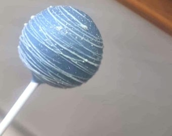 May include: A spherical cake pop with a smooth, matte slate blue coating, decorated with thin, swirling white icing drizzles and a light dusting of fine, shimmering edible glitter, attached to a plain white paper stick.