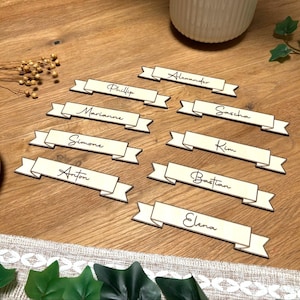 May include: Wooden place cards, each shaped like a banner with a light cream centre and dark brown edges. Names are written in elegant script, including Alexander, Phillip, Marianne, Simone, Anton, Sascha, Kim, Bastian, and Elena.