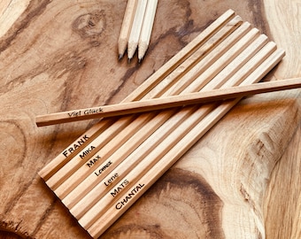 Puede incluir: Un conjunto de lápices de madera con nombres grabados. Los lápices están dispuestos sobre una superficie de madera. El texto en los lápices dice: "Viel Glück", "FRANK", "MIKA", "MAX", "Lennox", "Lene", "MATS" y "CHANTAL".