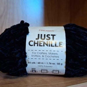 May include: A dark blue chenille yarn ball with a white label that reads "JUST CHENILLE" and "For Crafters, Makers, Knitters, & Crocheters." The label also indicates 65 yards, 1.76 oz, and 100% polyester.