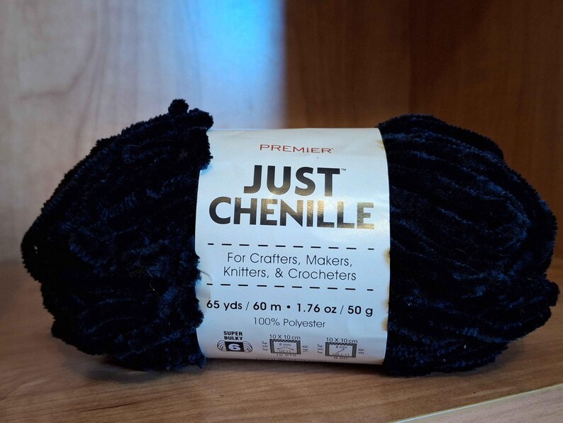 May include: A dark blue chenille yarn ball with a white label that reads "JUST CHENILLE" and "For Crafters, Makers, Knitters, & Crocheters." The label also indicates 65 yards, 1.76 oz, and 100% polyester.