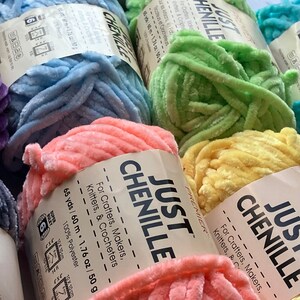 May include: Close-up of several skeins of Just Chenille yarn in various colors, including purple, blue, green, pink, and yellow. Each skein is labeled with the brand name and details for crafters, knitters, and crocheters.