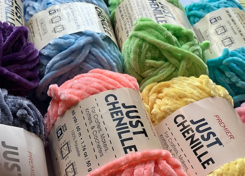 May include: Close-up of several skeins of Just Chenille yarn in various colors, including purple, blue, green, pink, and yellow. Each skein is labeled with the brand name and details for crafters, knitters, and crocheters.