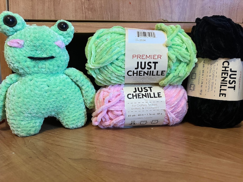 May include: A plush, mint green frog toy with black eyes and pink cheeks sits next to three skeins of chenille yarn in green, pink, and black. The yarn labels read "JUST CHENILLE" and indicate 65 yards (59.4 meters) of 100% polyester.