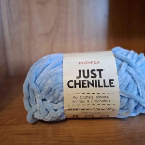 May include: A light blue chenille yarn ball with a label that reads "JUST CHENILLE" and "For Crafters, Makers, Knitters, & Crocheters." The label also indicates 65 yards, 1.76 oz, and 100% polyester.
