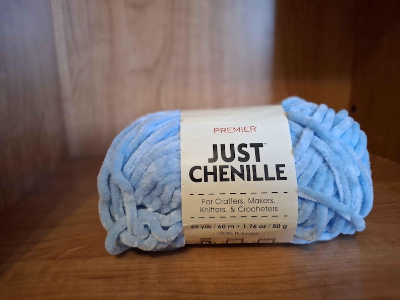 May include: A light blue chenille yarn ball with a label that reads "JUST CHENILLE" and "For Crafters, Makers, Knitters, & Crocheters." The label also indicates 65 yards, 1.76 oz, and 100% polyester.