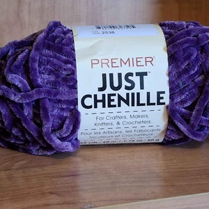 May include: A skein of Premier Just Chenille yarn in a deep purple hue. The label reads "For Crafters, Makers, Knitters, & Crocheters." The yarn is 48 yards / 40 m and weighs 1.76 oz / 50 g. The yarn is on a wooden surface.