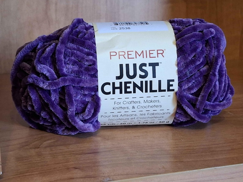 May include: A skein of Premier Just Chenille yarn in a deep purple hue. The label reads "For Crafters, Makers, Knitters, & Crocheters." The yarn is 48 yards / 40 m and weighs 1.76 oz / 50 g. The yarn is on a wooden surface.