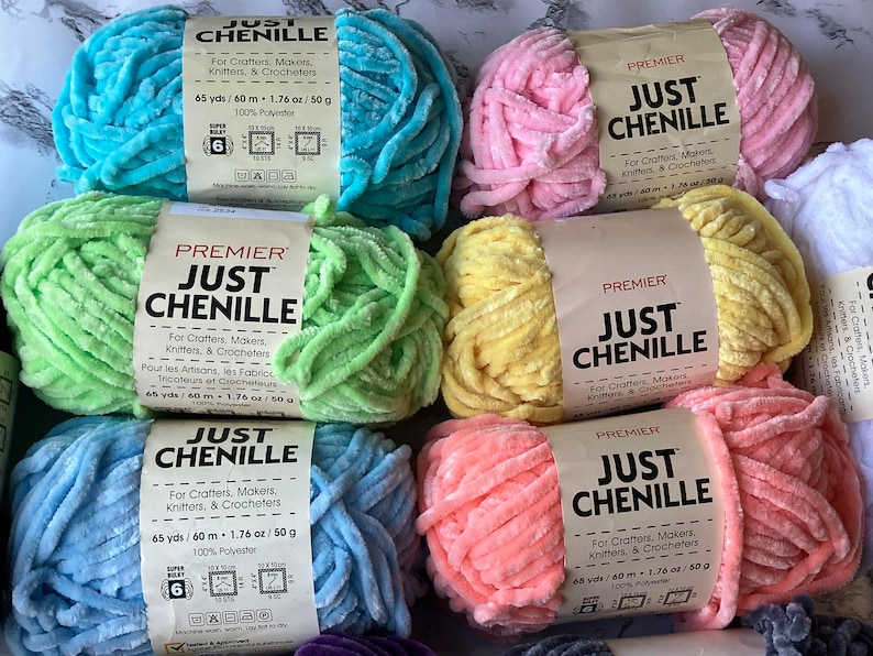 May include: A collection of Premier Just Chenille yarn skeins in various pastel colors, including blue, pink, green, and yellow. Each skein is labeled with text for crafters, knitters, and crocheters. The yarn is 65 yards/60 meters and made of 100% polyester.