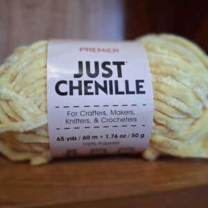 May include: A skein of Premier Just Chenille yarn in a soft yellow color. The label reads "For Crafters, Makers, Knitters, & Crocheters." The yarn is 65 yards / 60 meters and 1.76 oz / 50 g of 100% polyester.