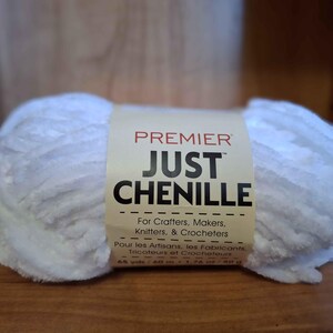 May include: A white chenille yarn ball with a cream-colored label. The label reads "PREMIER JUST CHENILLE" and includes text for crafters, makers, knitters, and crocheters. The yarn is 65 yards / 60 m.