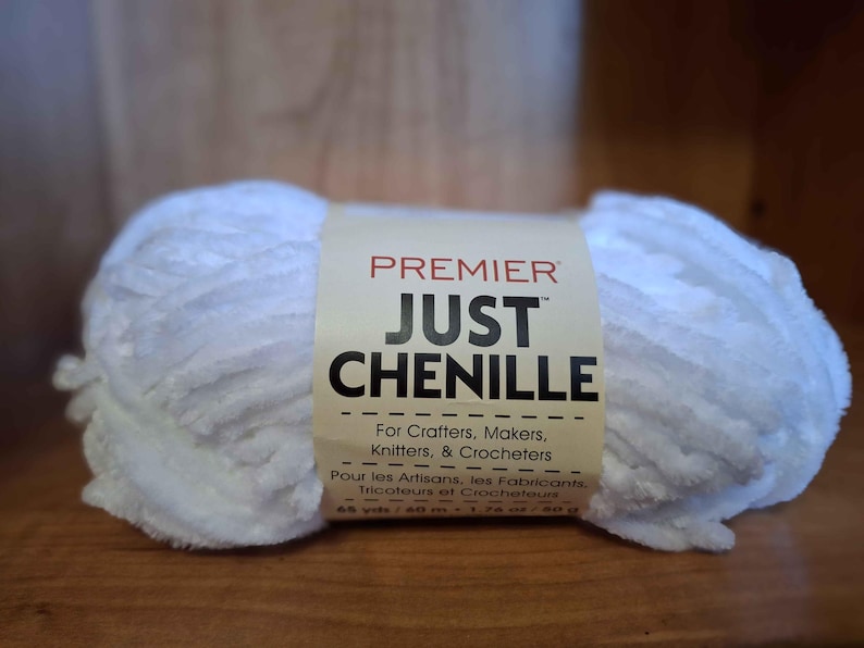 May include: A white chenille yarn ball with a cream-colored label. The label reads "PREMIER JUST CHENILLE" and includes text for crafters, makers, knitters, and crocheters. The yarn is 65 yards / 60 m.