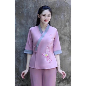 May include: A woman wearing a light pink two-piece outfit with a floral design. The top has a grey trim and a mandarin collar. The pants are loose fitting.