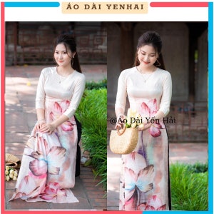 Puede incluir: Dos mujeres con camisas blancas de manga larga con una falda de estampado floral. La falda tiene un estampado floral rosa, blanco y azul. Las mujeres están de pie frente a un arbusto verde. El texto "@Ao Dai Yen Hai" es visible en la imagen.