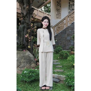 May include: A woman wearing a beige two-piece outfit with a brown collar and buttons. The top has a small embroidered design on the front. She is standing in front of a stone wall and a tree.