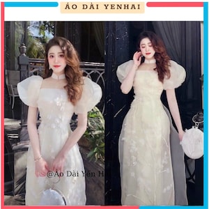 May include: A white traditional Vietnamese dress with puffed sleeves and floral embroidery. The dress has a high neckline and a fitted bodice. The dress is worn by a woman with long brown hair. The dress is called "Áo Dài Yến Hai" and the text "@Áo Dài Yến Hai" is visible in the image.