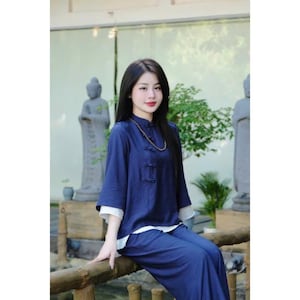 May include: A woman wearing a dark blue traditional Chinese-style top with a white collar and matching trousers. She is sitting on a wooden railing with a background of greenery and statues.