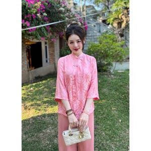 May include: A woman wearing a pink, floral patterned blouse with a mandarin collar and a matching pink skirt. She is holding a white handbag with a gold clasp.