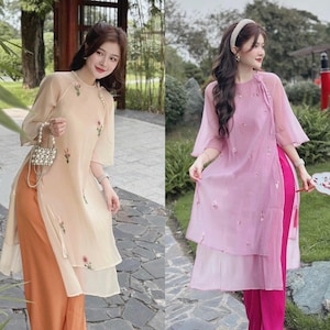 May include: Two women wearing traditional Vietnamese dresses, one in a light pink dress with a pink and white floral pattern and the other in a cream dress with a cream and pink floral pattern. Both dresses have a side slit and are paired with matching pants.