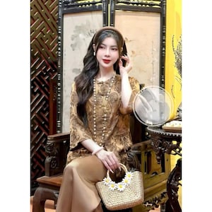 Puede incluir: Una mujer con una blusa de estampado marrón y dorado con un collar de perlas y una diadema marrón. Lleva un bolso de paja con flores blancas de margarita.