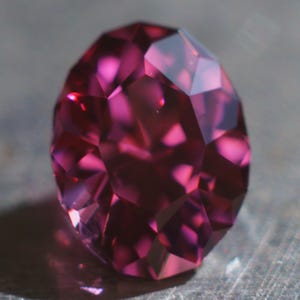 Puede incluir: Una piedra preciosa rosa intenso con un corte facetado. La piedra preciosa tiene forma ovalada y un alto pulido.