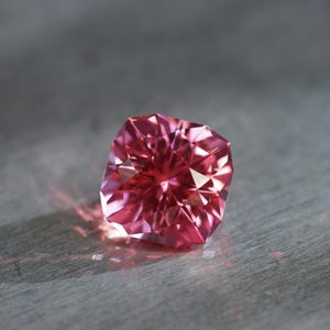 Peut inclure: Une pierre pr&eacute;cieuse rose avec une taille carr&eacute;e et de nombreuses facettes. La pierre pr&eacute;cieuse est pos&eacute;e sur une surface grise.