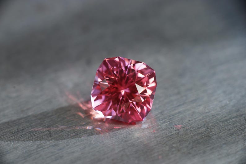 Peut inclure: Une pierre pr&eacute;cieuse rose avec une taille carr&eacute;e et de nombreuses facettes. La pierre pr&eacute;cieuse est pos&eacute;e sur une surface grise.