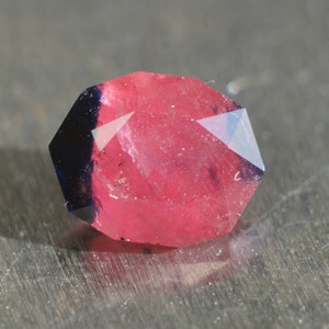 Puede incluir: Una gema facetada, predominantemente rosa con una sección azul oscuro. La gema tiene múltiples superficies planas que reflejan la luz. La piedra se muestra sobre un fondo neutro, resaltando su color y forma.