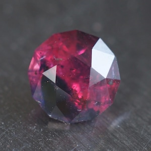 May include: A faceted gemstone in a deep magenta colour. The gem has multiple flat surfaces, creating a geometric shape. The stone's colour varies from dark red to a lighter pink, with some areas appearing almost black.