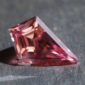 May include: A pink gemstone with a faceted, triangular shape. The gem is cut in a unique style with multiple facets.