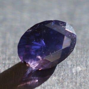 May include: A faceted, oval-shaped gemstone with a deep indigo hue. The gem displays a complex pattern of internal reflections and facets, with a shadow cast to the lower left. The surface has some minor imperfections.