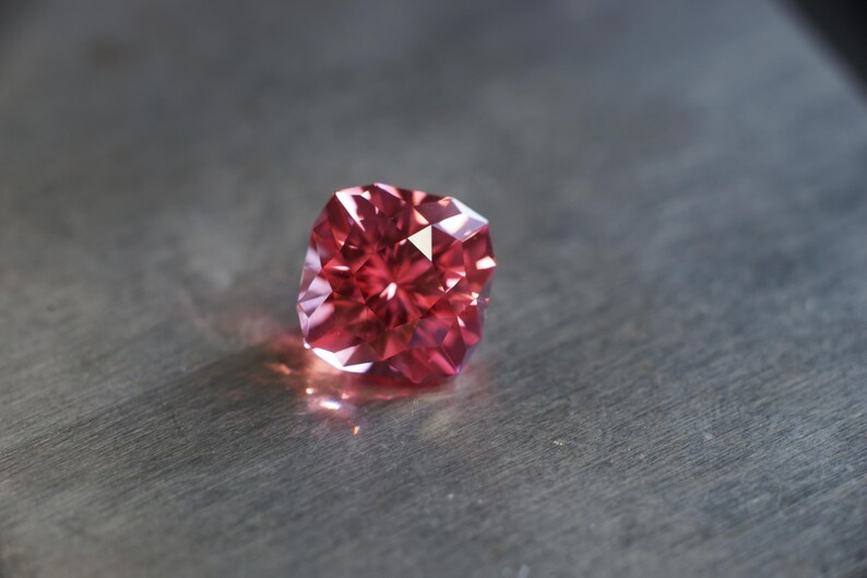 Peut inclure: Une pierre pr&eacute;cieuse rouge de forme carr&eacute;e avec de nombreuses facettes. La pierre pr&eacute;cieuse est pos&eacute;e sur une surface grise.