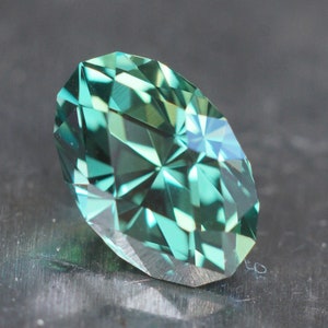 May include: A faceted, oval-shaped gemstone with a teal and green color gradient. The gem's surface reflects light, creating a sparkling effect. The gemstone is set against a dark, textured background, highlighting its brilliance.
