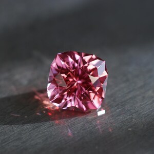 Peut inclure: Une pierre pr&eacute;cieuse rose avec une taille carr&eacute;e et de nombreuses facettes. La pierre pr&eacute;cieuse est pos&eacute;e sur une surface gris fonc&eacute;.