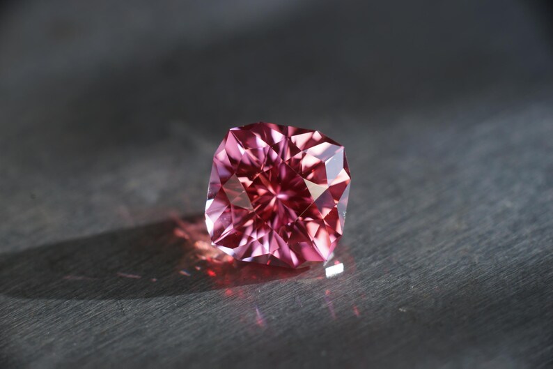 Peut inclure: Une pierre pr&eacute;cieuse rose avec une taille carr&eacute;e et de nombreuses facettes. La pierre pr&eacute;cieuse est pos&eacute;e sur une surface gris fonc&eacute;.