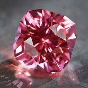 May include: A pink gemstone with a faceted cut, reflecting light on a gray surface.