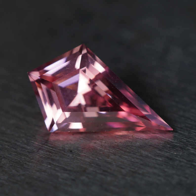 May include: A pink, faceted gemstone with a triangular shape. The gemstone is cut in a unique, geometric style and has a high level of clarity.