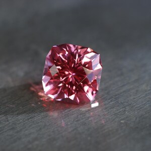 Peut inclure: Une pierre pr&eacute;cieuse rose &agrave; facettes, pr&eacute;sentant un motif en &eacute;toile. La pierre pr&eacute;cieuse est plac&eacute;e sur un fond gris.