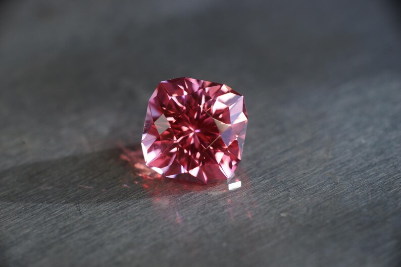Peut inclure: Une pierre pr&eacute;cieuse rose &agrave; facettes, pr&eacute;sentant un motif en &eacute;toile. La pierre pr&eacute;cieuse est plac&eacute;e sur un fond gris.