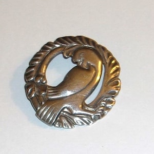 May include: A silver brooch in the shape of a bird with its wings spread out. The bird is surrounded by a circle of leaves.