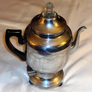 May include: A vintage silver-toned electric coffee percolator with a black handle and a glass top. The percolator has a decorative etched design around the middle and sits on a round base. A classic kitchen appliance.